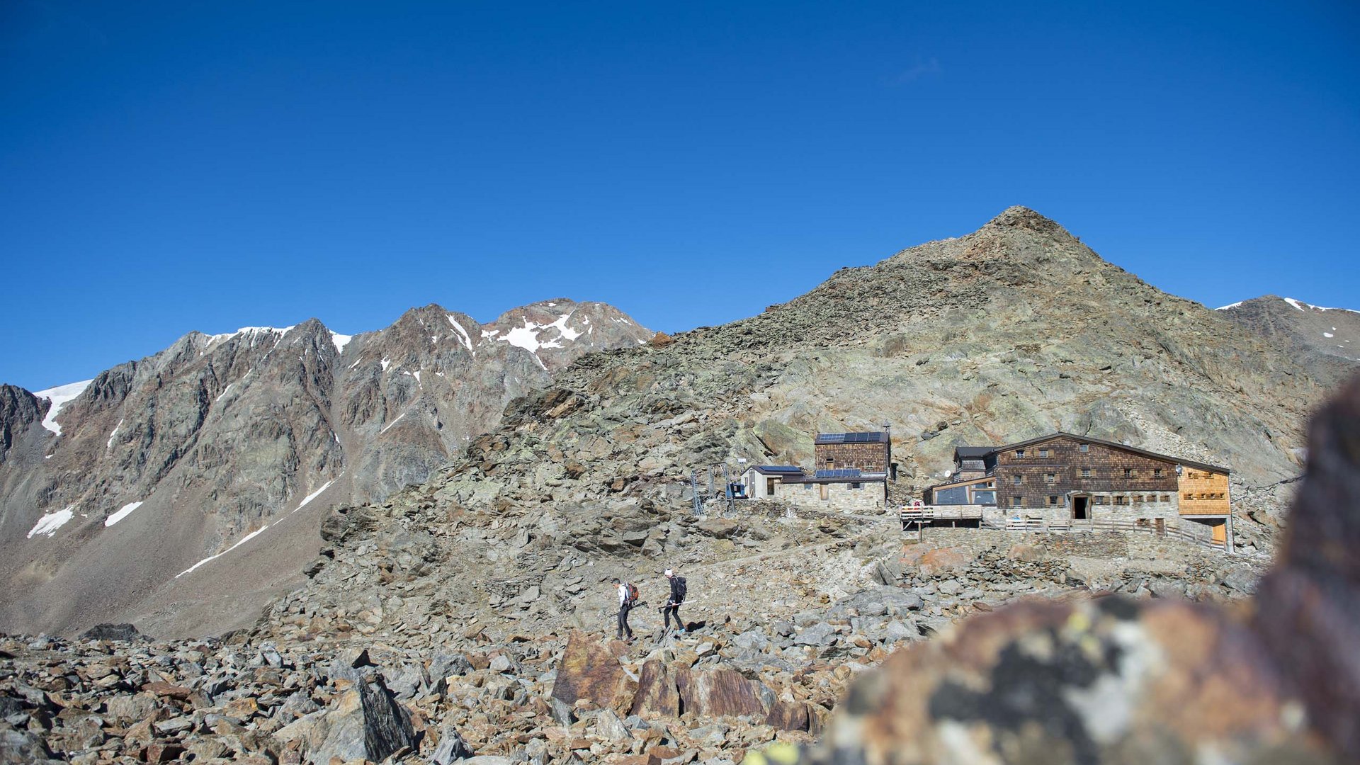 Our favourite tours – incl. the walk up to Similaun hut Hikers on rocky trail near mountain hut under clear blue sky