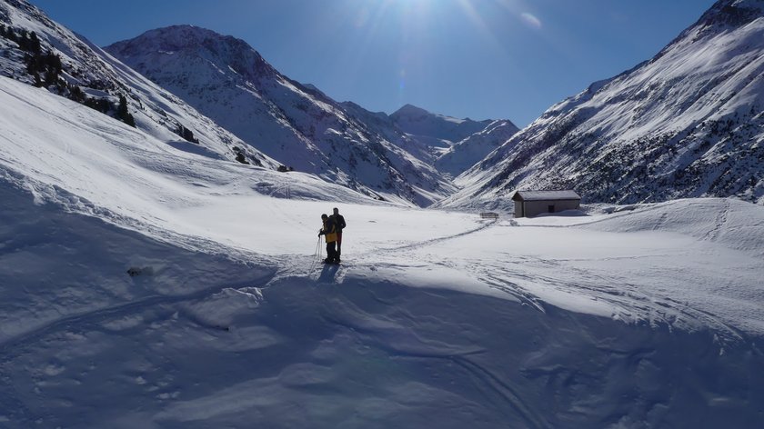 From Vent to the Ramolalm Two skiers in snowy valley with mountains and cabin under bright sunny sky
