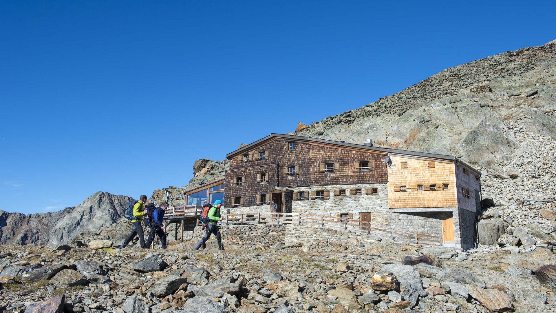 Alpinclub Similaun: durch die Liebe zum Berg vereint Drei Wanderer nähern sich einer Berghütte im felsigen alpinen Gelände bei klarem Himmel