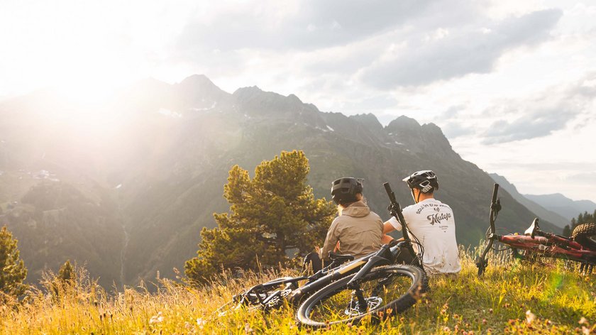 Mountainbiken im Ötztal – Trails für alle Niveaus Zwei Radfahrer sitzen auf einer Bergwiese und genießen den Ausblick bei Sonnenuntergang