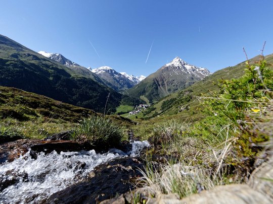Wandern in den Ötztaler Alpen: von Ihrem Hotel in die Berge Bergbach fließt durch grüne Alpenwiese mit schneebedeckten Gipfeln im Hintergrund