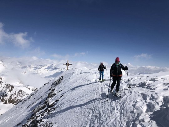 Backcountry skiing near Vent: start directly from our hotel Two skiers hiking on snowy mountain peak with cross under clear sky