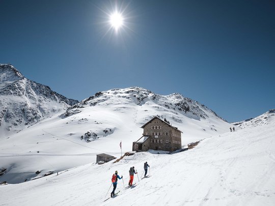 Backcountry skiing near Vent: start directly from our hotel Three skiers near a mountain hut in snowy alpine landscape under bright sun