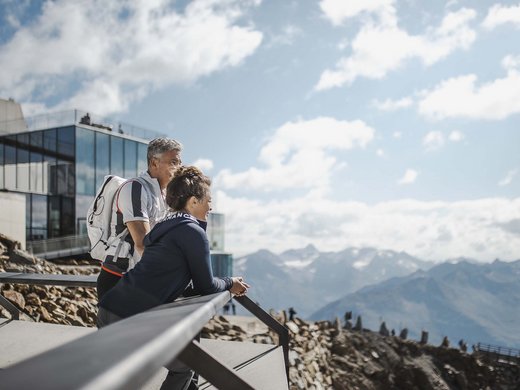A summer holiday in Ötztal: a paradise for mountain lovers Two hikers looking at mountain landscape from a terrace with modern buildings