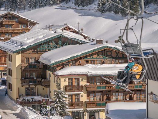 Werden Sie Teil des Teams unseres Hotels im Ötztal in Tirol! Zwei Skifahrer in einer Sesselliftkabine vor schneebedeckten Alpenhäusern