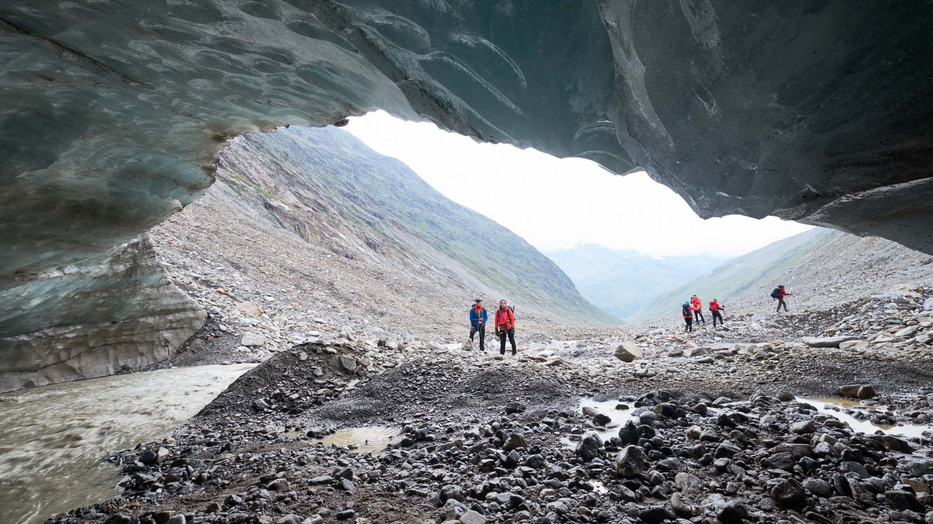 Our favourite tours – incl. the walk up to Similaun hut Hikers under an ice cave with rocky valley and river in the background